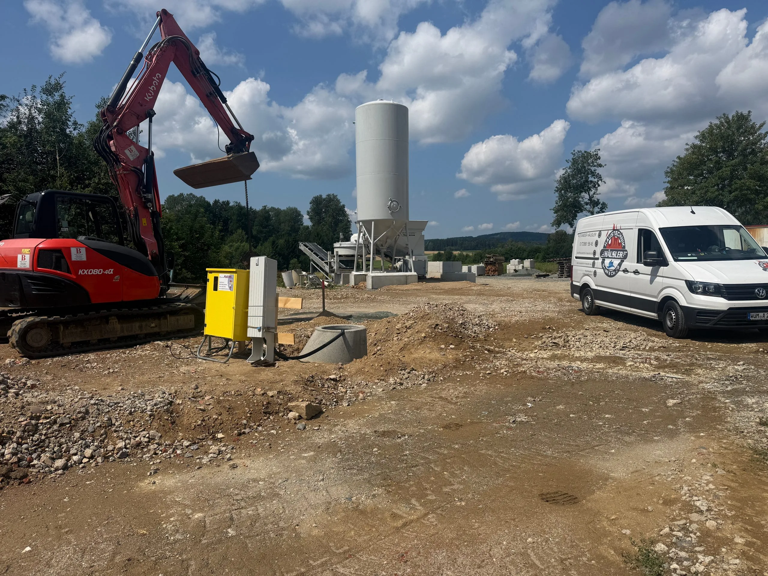 Baustelle mit Häusler-Haustechnik-Servicewagen rechts und einer mobilen Betontankstelle mit Silo mittig, daneben ein roter Kubota-Bagger – Wasserversorgung der Betontankstelle für Bagger & Verleih Bauer in Muschenried.