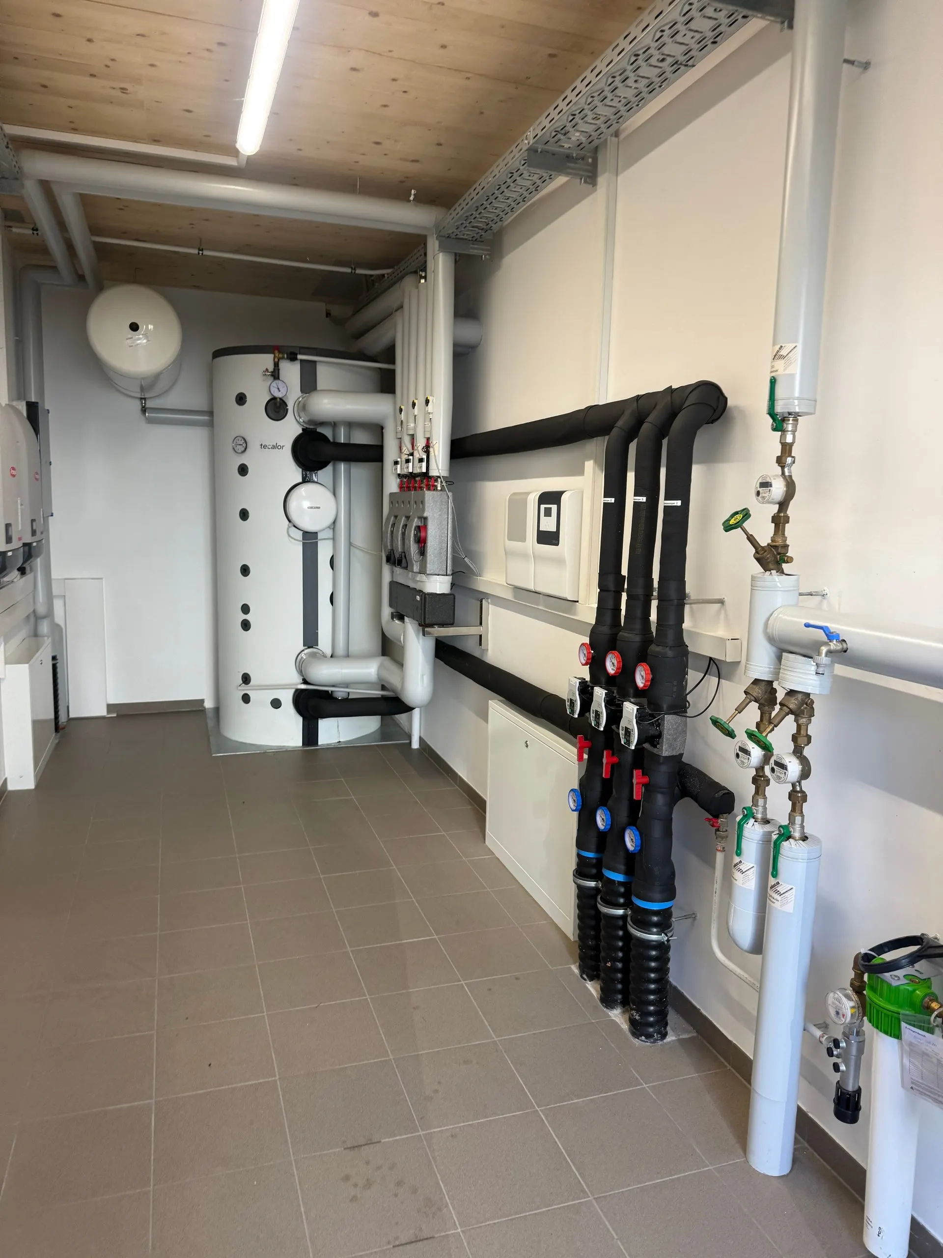 Innenansicht des Technikraums mit Tecalor-Pufferspeicher, Verteilern und gedämmten Vor- und Rücklaufleitungen der Wärmepumpenanlage.